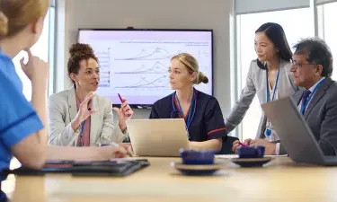 Healthcare administrators and professionals collaborating in a meeting room with a presentation displayed on a screen, discussing data and strategies.